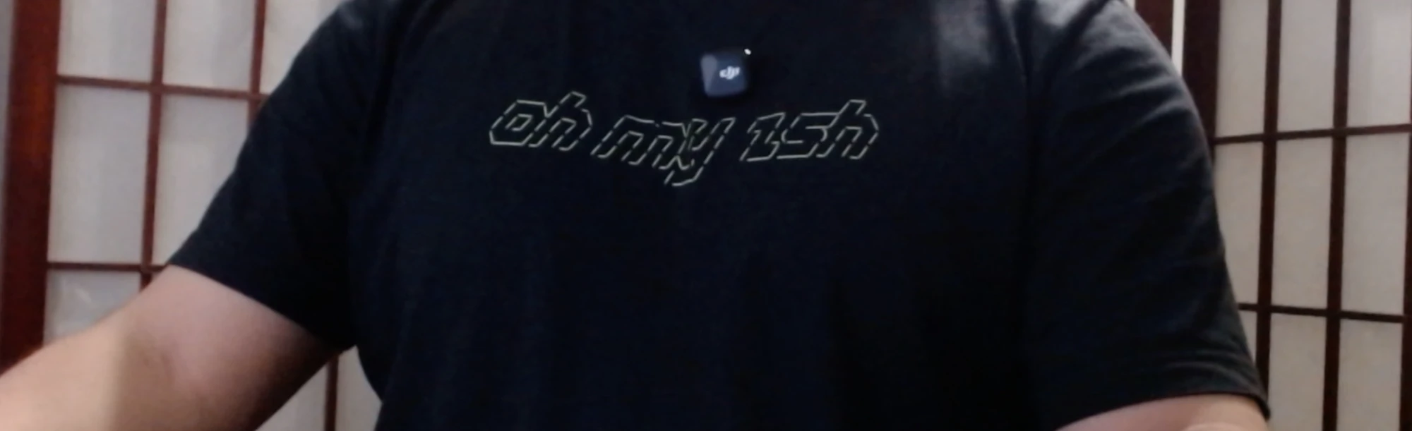Luis in a black t-shirt printed with “oh my zsh” in outlined, slanted lettering, a small DJI wireless microphone clipped to the collar, in front of a shoji screen
