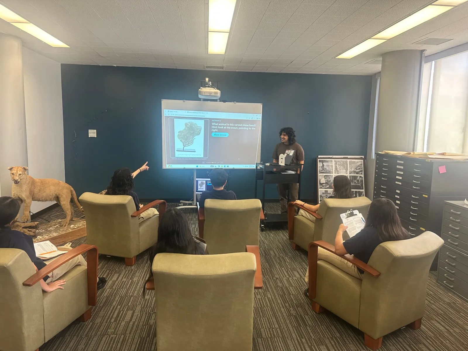 Station 7 in the Special Collections reading room: 7th grade students in armchairs face a projected slide asking “What animal is this carved stone head?”, one student pointing at the screen, while a taxidermy cougar stands to the left and the presenter smiles beside a cart marked with a number 7