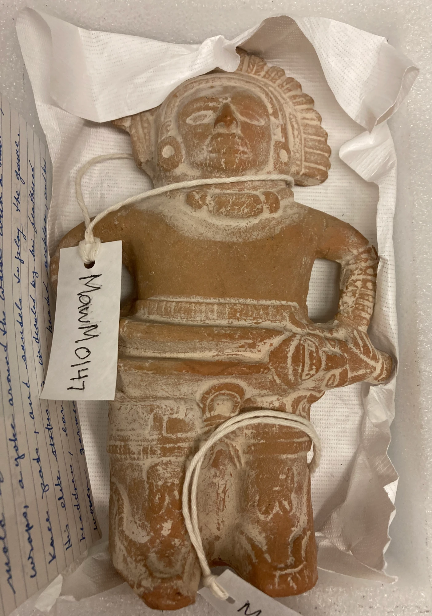 A clay figure of a Mesoamerican ballplayer with a feathered headdress and a thick yoke around the waist, resting in tissue paper in a storage box with a handwritten tag reading MawM0147