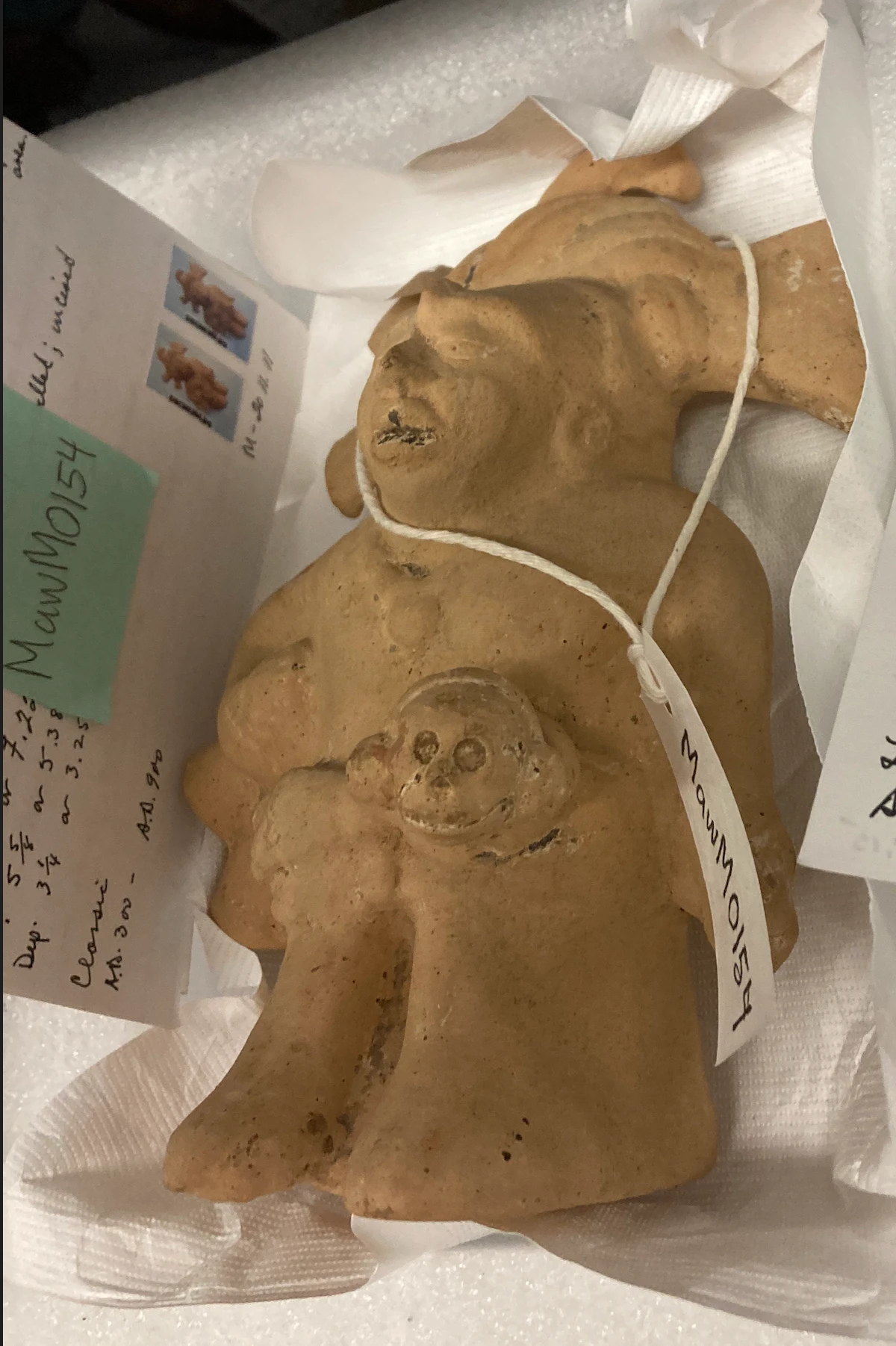The same clay figure seen from the front, seated and holding a small round-eyed face in its lap.