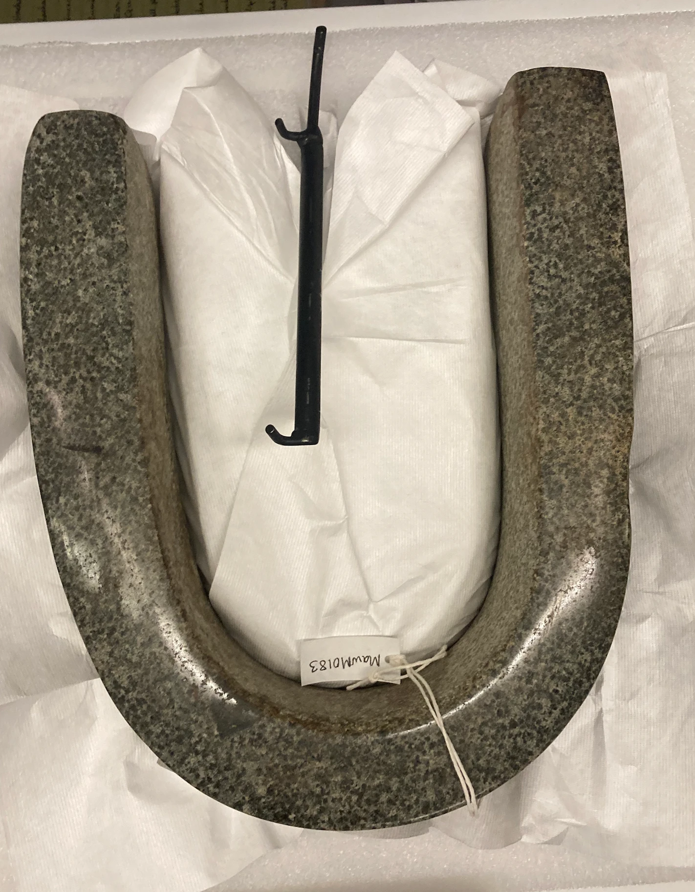 A heavy U-shaped stone yoke resting in a storage box, tagged MawM0183.
